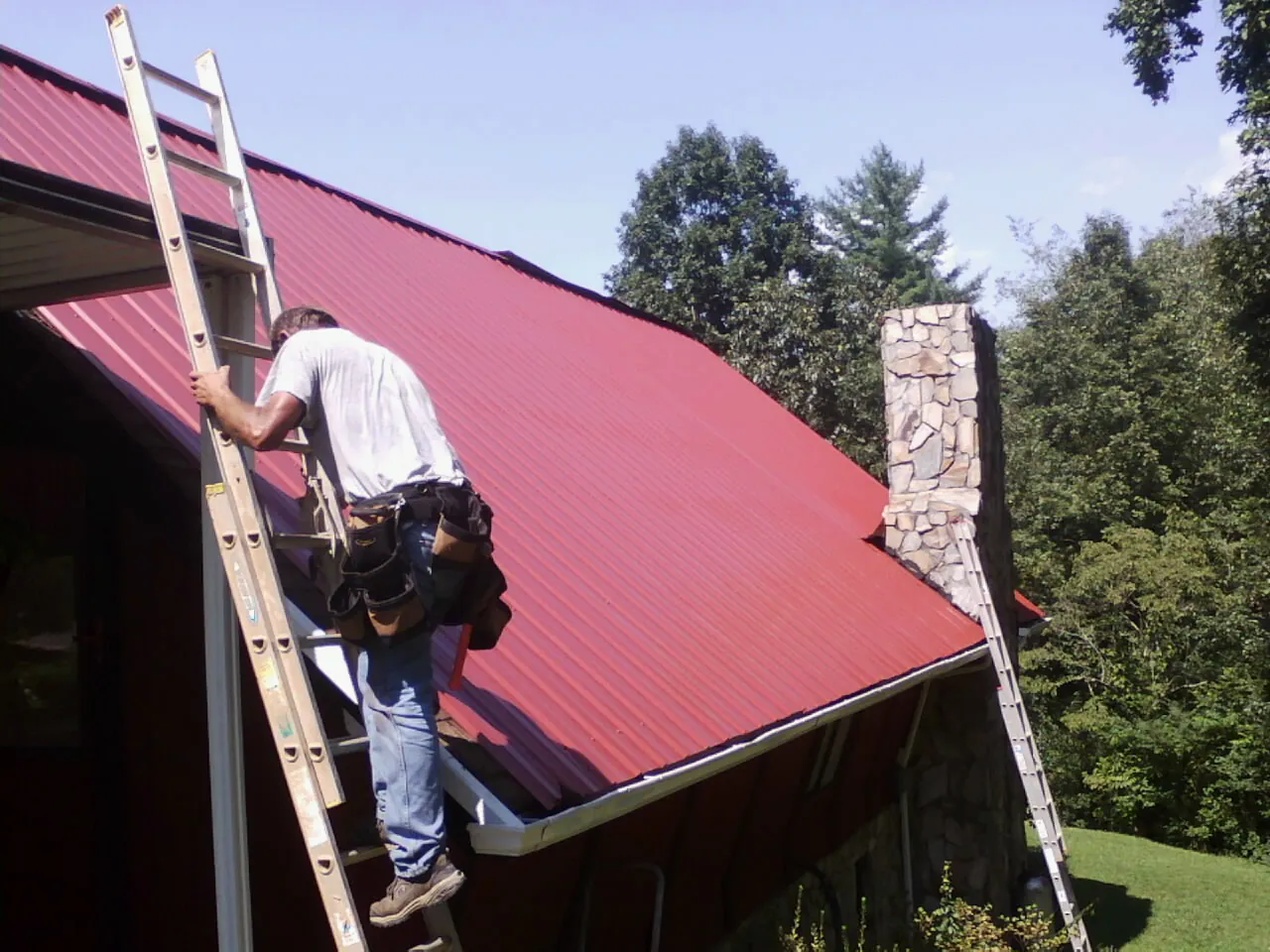 Roofer on a ladder beside a red metal roof and stone chimney — Soffit & Fascia Repair by Roofing Brothers in Perkinston